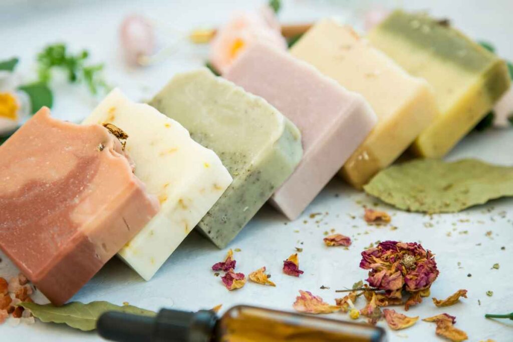 A selection of five handmade soap bars in various colors arranged diagonally on a light surface, surrounded by dried flowers, leaves, and a small dropper bottle.