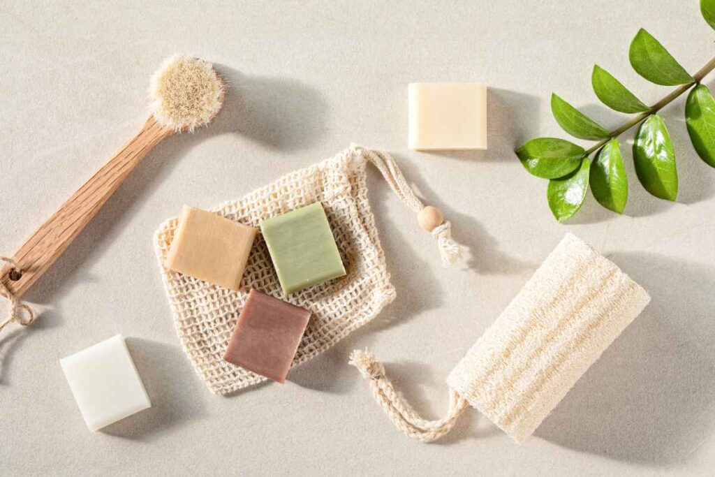 An assortment of natural soap bars in different colors, a wooden bath brush, a loofah, and a green leaf on a neutral background.