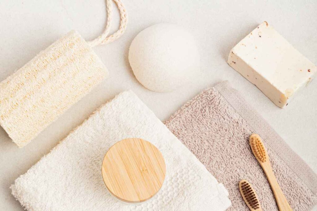 A flat lay of eco-friendly bathroom accessories including a loofah, towels, wooden toothbrushes, a soap bar, and a round facial sponge, all in soft neutral tones.