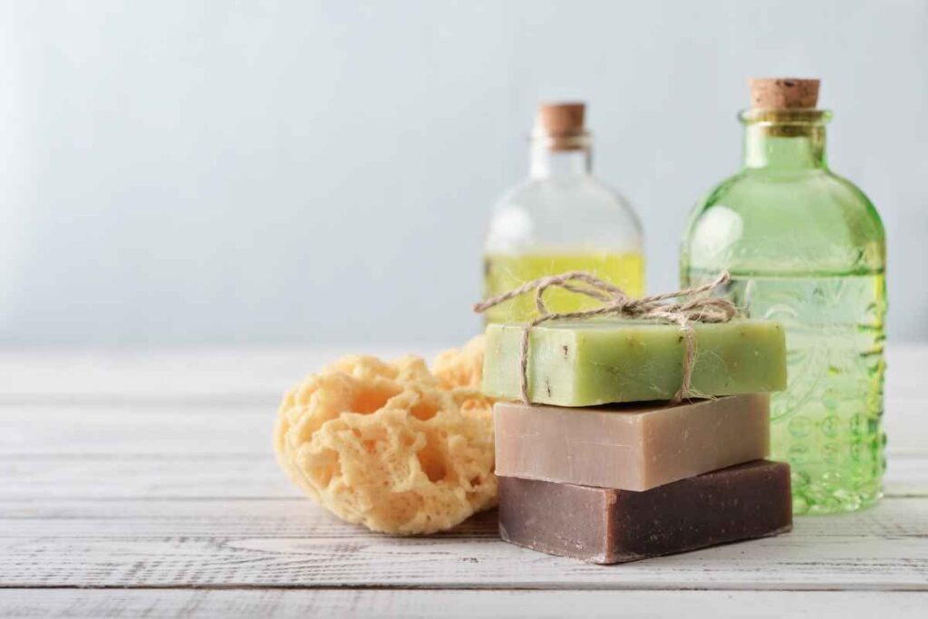 A stack of three handmade soap bars in green, brown, and beige, tied with twine, next to a natural sea sponge and glass bottles on a wooden surface.