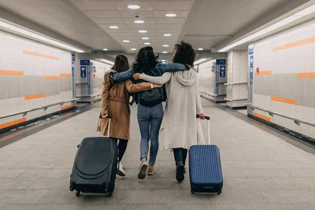 Three friends embracing each other in camaraderie, walking through a subway station as houseguests during travel, pulling along their suitcases and symbolizing the journey and connection inherent in travel.