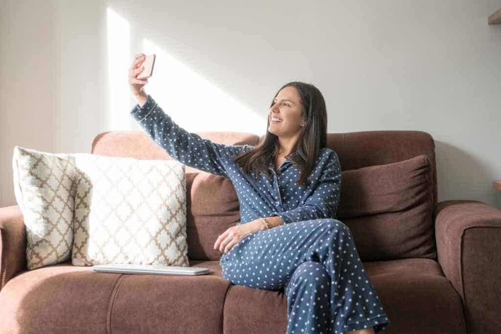 A houseguest during travel relaxes in a polka-dot pajama set, capturing a cheerful moment with a selfie, while seated on a brown couch adorned with decorative pillows, embodying a carefree and respectful temporary residence.