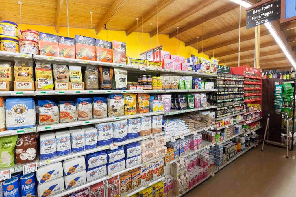 Being a Good Houseguest While Traveling can include contributing to the household, depicted by a grocery store shelf stocked with various baking ingredients like flour and sugar, under a sign labeling 'Bake items', in a well-lit, organized shopping environment.