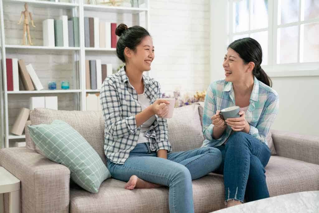 Two friends share a light-hearted moment over coffee in a brightly-lit living room, embodying the key aspects of Being a Good Houseguest While Traveling with their comfortable and engaged interaction in a homely setting