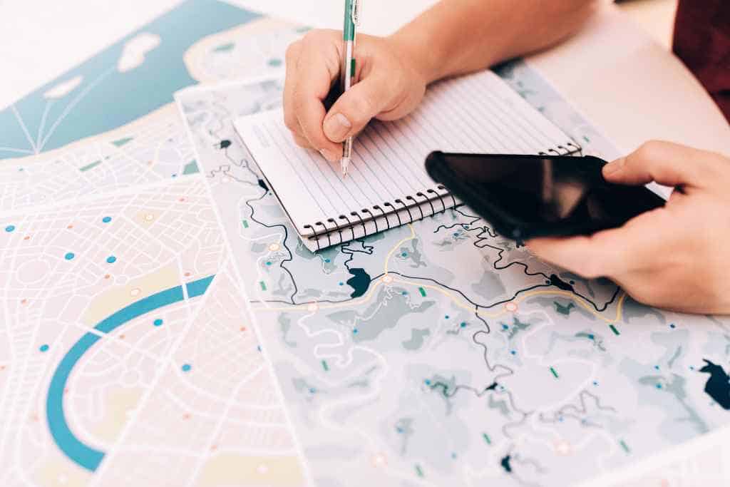 Houseguest during travel is engrossed in planning adventures, with a hand holding a pen over a spiral notebook, another hand navigating a smartphone, all laid out next to a detailed city map, highlighting organization skills.