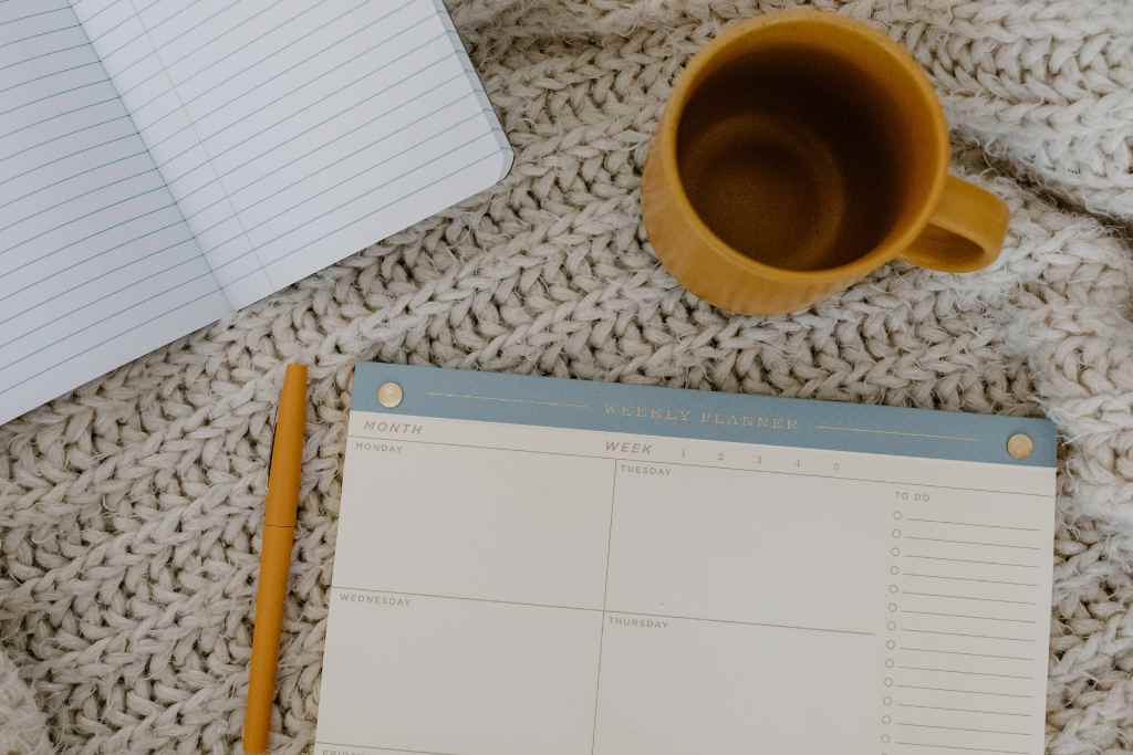 Organizational skills are key to Being a Good Houseguest While Traveling, depicted by a neatly laid out weekly planner with a 'MONTH' and 'WEEK' section visible, a blank notebook for additional notes, a wooden pencil, all complemented by a comforting knit blanket and a mug of coffee.