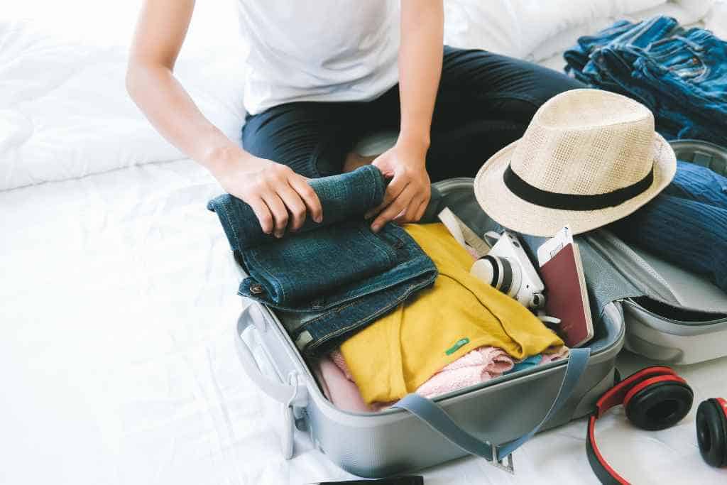 The essence of a houseguest during travel captured in the act of meticulously rolling denim jeans and packing bright yellow and blue clothing, alongside essential electronic gadgets and a stylish straw hat, into a spacious gray suitcase on a white bedspread.