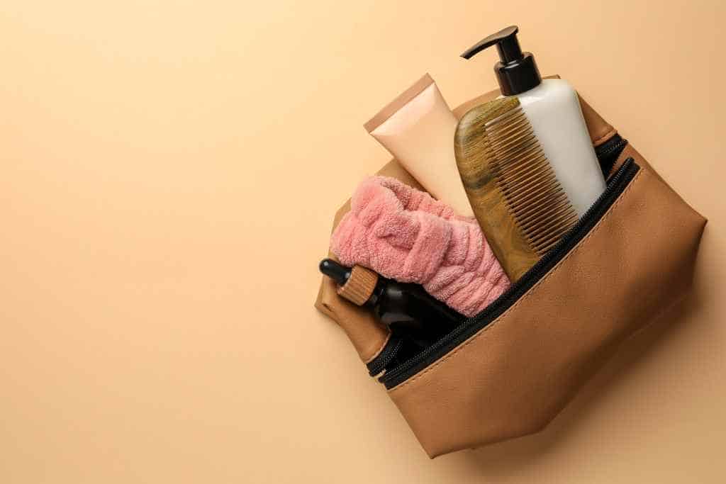 A considerate houseguest during travel packs a brown toiletry bag with personal care essentials, featuring bottles, a comb, and a soft towel, arranged on a muted beige background, signifying neatness and preparedness.