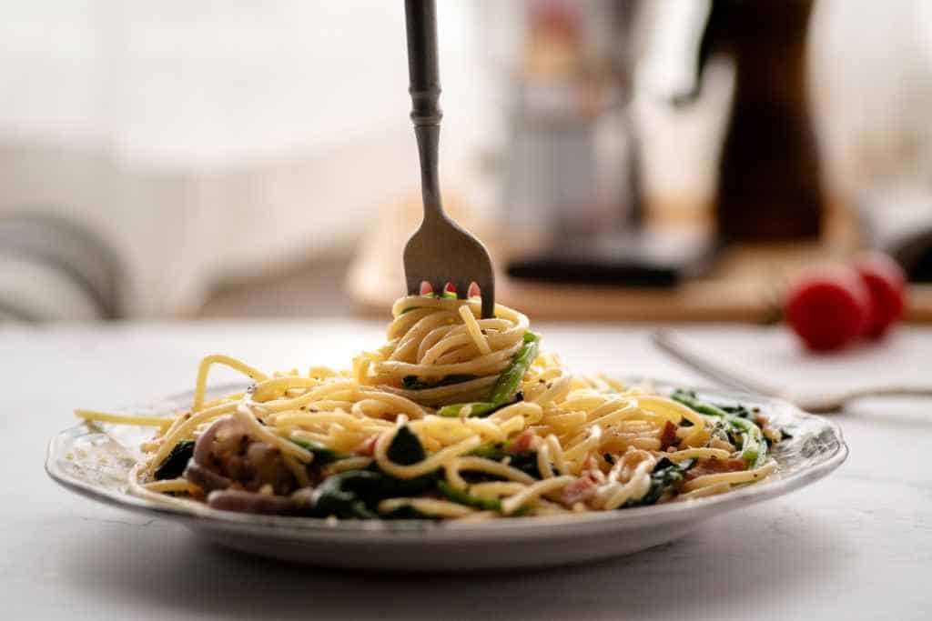 Sharing a homemade meal is part of Being a Good Houseguest While Traveling, as seen in this delectable dish of spaghetti aglio e olio adorned with fresh greens, presented on a ceramic plate set on a white table, ready to be enjoyed.