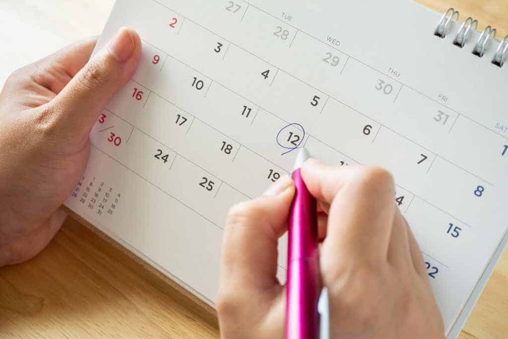 A houseguest during travel demonstrates thoughtful planning by marking a specific date with a pink highlighter on a desk calendar, ensuring they are considerate of their host's schedule and important events.