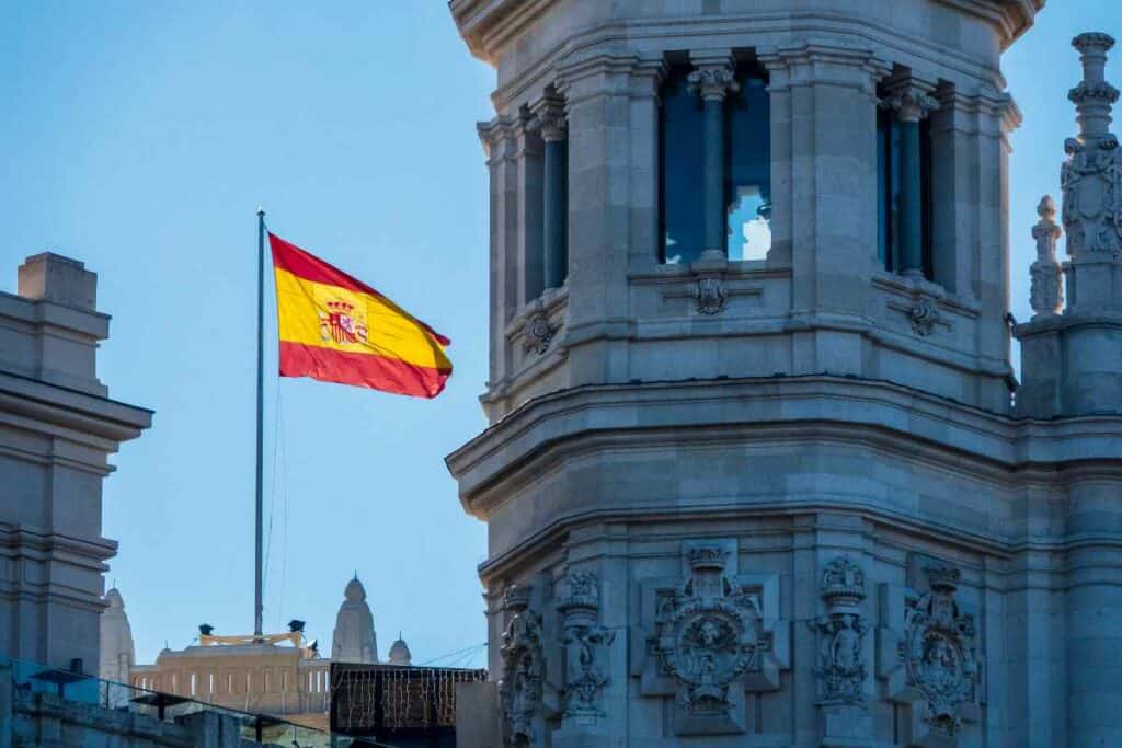 The Spanish flag waving in front of a historic building with ornate architectural details and columns under a clear blue sky.