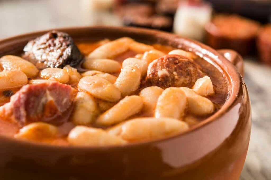 A close-up of a traditional Spanish stew featuring white beans, chorizo, and chunks of meat in a rustic clay bowl.
