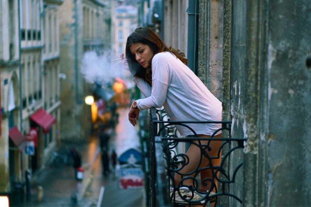 A woman leaning on a wrought-iron balcony railing, exhaling smoke while looking down at a bustling, narrow street below.