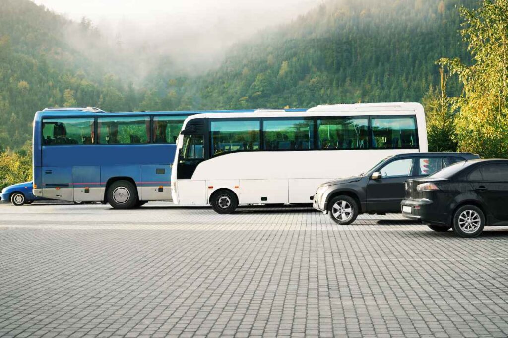 Two large buses, one blue and one white, are parked next to cars in a lot with a backdrop of misty green mountains.