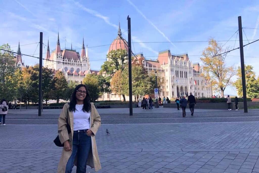 A smiling woman stands in front of the Hungarian Parliament Building in Budapest, with its iconic Gothic Revival architecture and domes, under a bright blue sky. This highlights the grandeur of the city and answers "Is Budapest Worth Visiting" with a resounding yes.