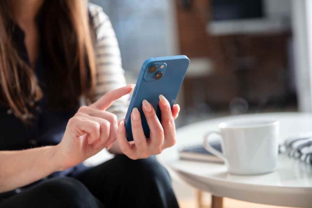 A woman seated at a table, using a blue smartphone with a white coffee mug and notebook in the background.