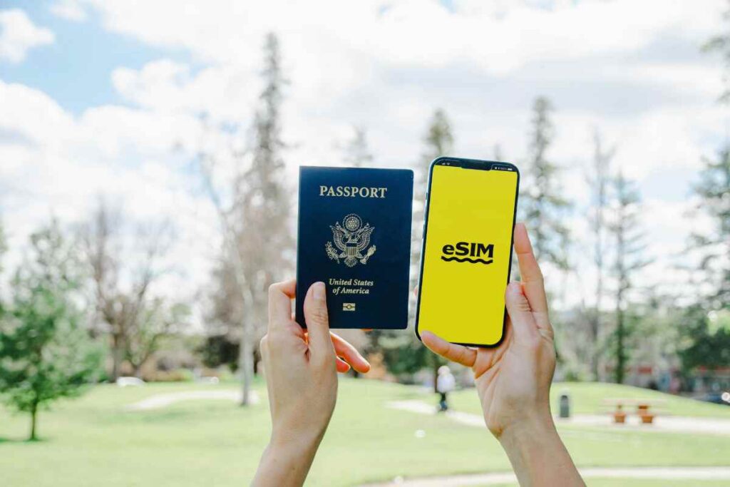 Hands holding a United States passport an a smartphone with an eSIM app open, set against a green park background on a sunny day.