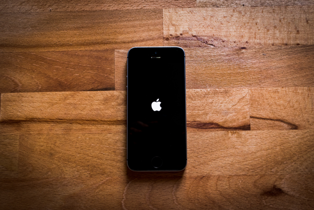 A black iPhone lying on a wooden surface, displaying the Apple logo on its screen as it powers on. The warm wood grain contrasts with the sleek design of the phone.