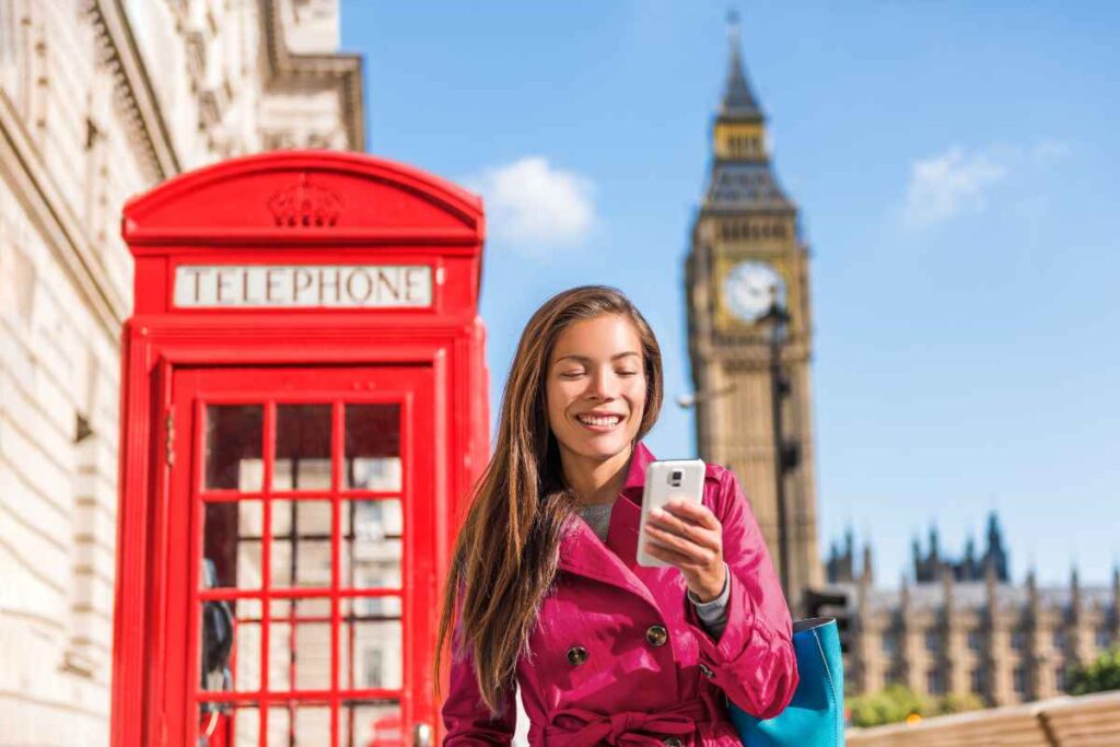  woman wearing a pink coat smiles while using a smartphone, standing near a red telephone booth with Big Ben and blue skies in the background.