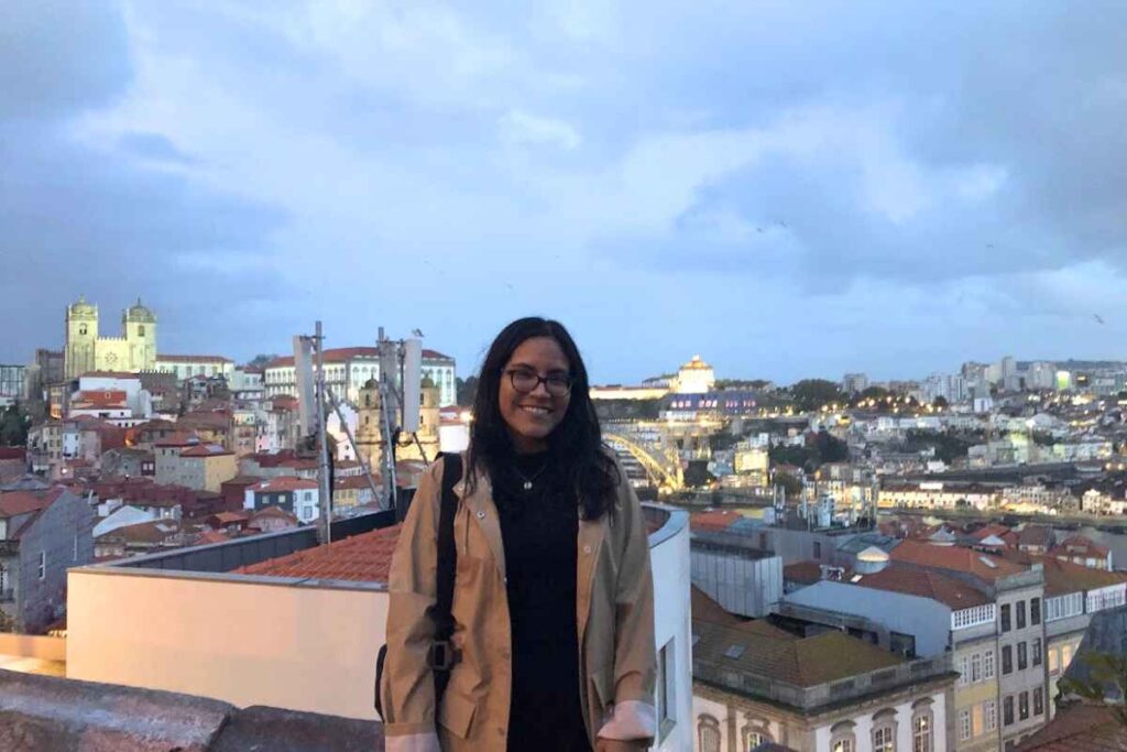 A solo traveler poses on a rooftop at dusk, with Porto's cityscape and historic cathedral illuminated in the background. The view highlights the charm of Porto, Portugal, answering the question, "Is Porto Portugal worth visiting?"