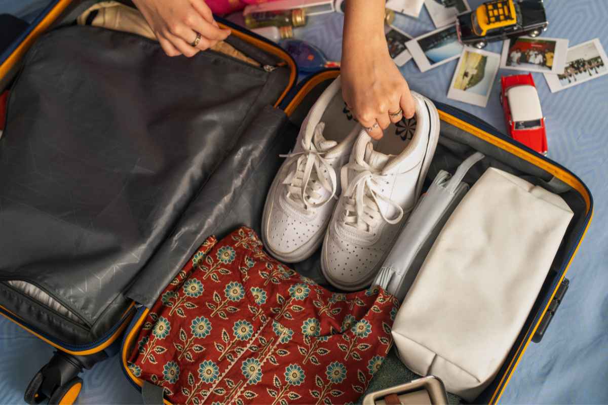 A traveler packs a suitcase, placing a pair of white sneakers neatly inside. The suitcase contains a red patterned fabric item, a white toiletry bag, and a folded umbrella. Scattered around the suitcase on a blue surface are small travel souvenirs, including toy cars and printed photographs. This image represents organized packing and selecting the Best Shoes for Europe Travel, ensuring comfort and style for walking long distances.